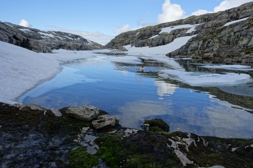 Felsen, dazwischen ein blauer Bergsee mit Schnee am Rand, darin eine kleine Schwimmerin