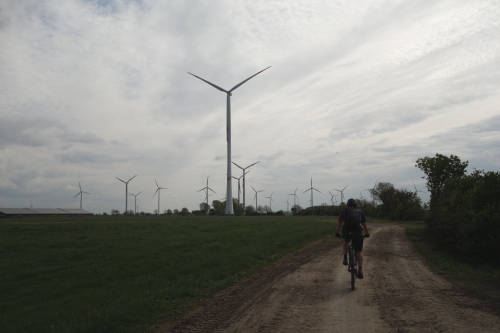 Radfahrer auf einem sandigen Feldweg unter stürmischem Himmel, im Hintergrund Windkraftanlagen