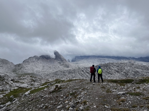 Zwei Wanderer mit bunten Rucksäcken vor grauer Steinwüste unter grauem Himmel